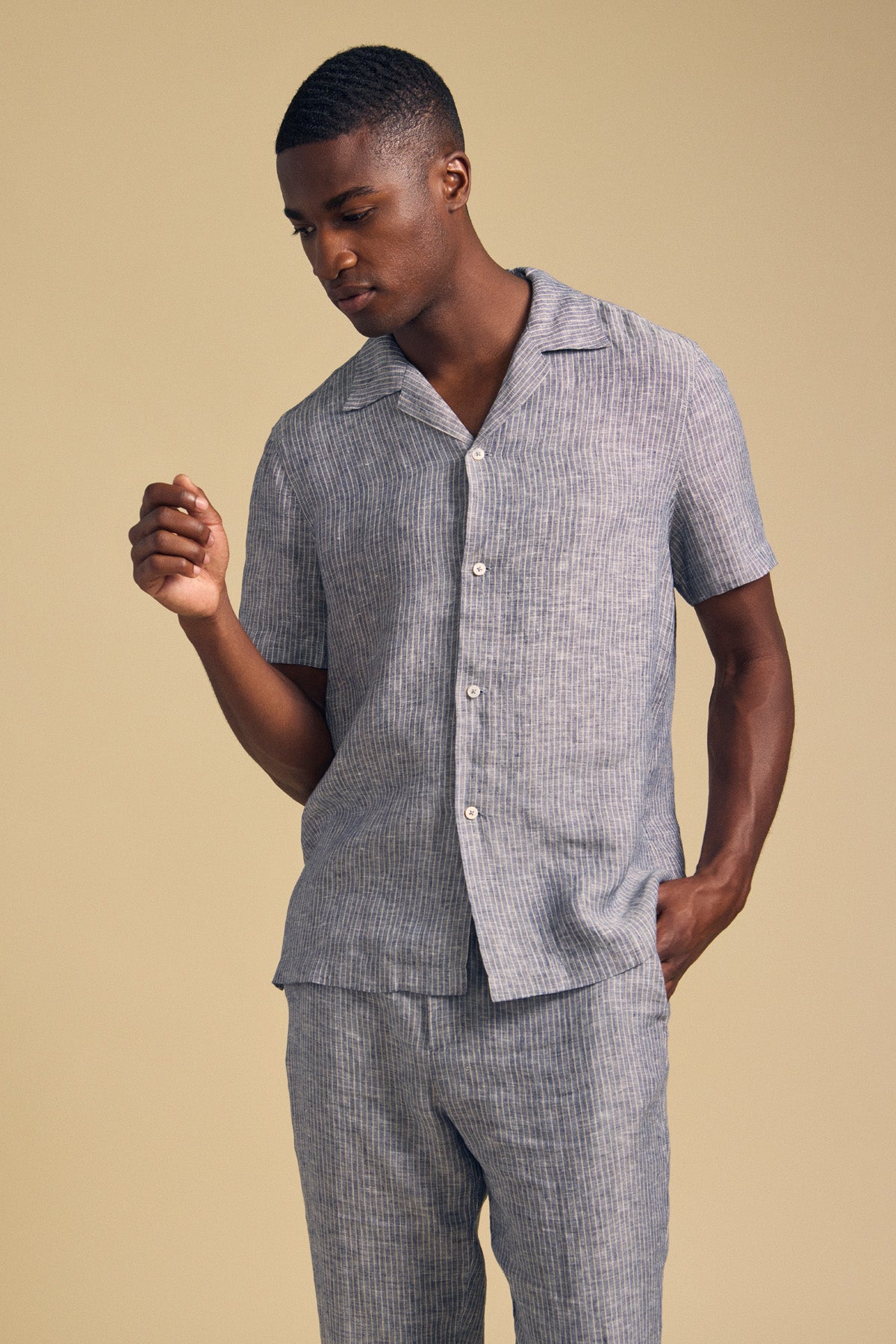 A man wearing the JULIUS LINEN SHIRT by Velvet by Graham & Spencer, a short-sleeve, button-up striped linen shirt with matching pants, stands against a beige background.