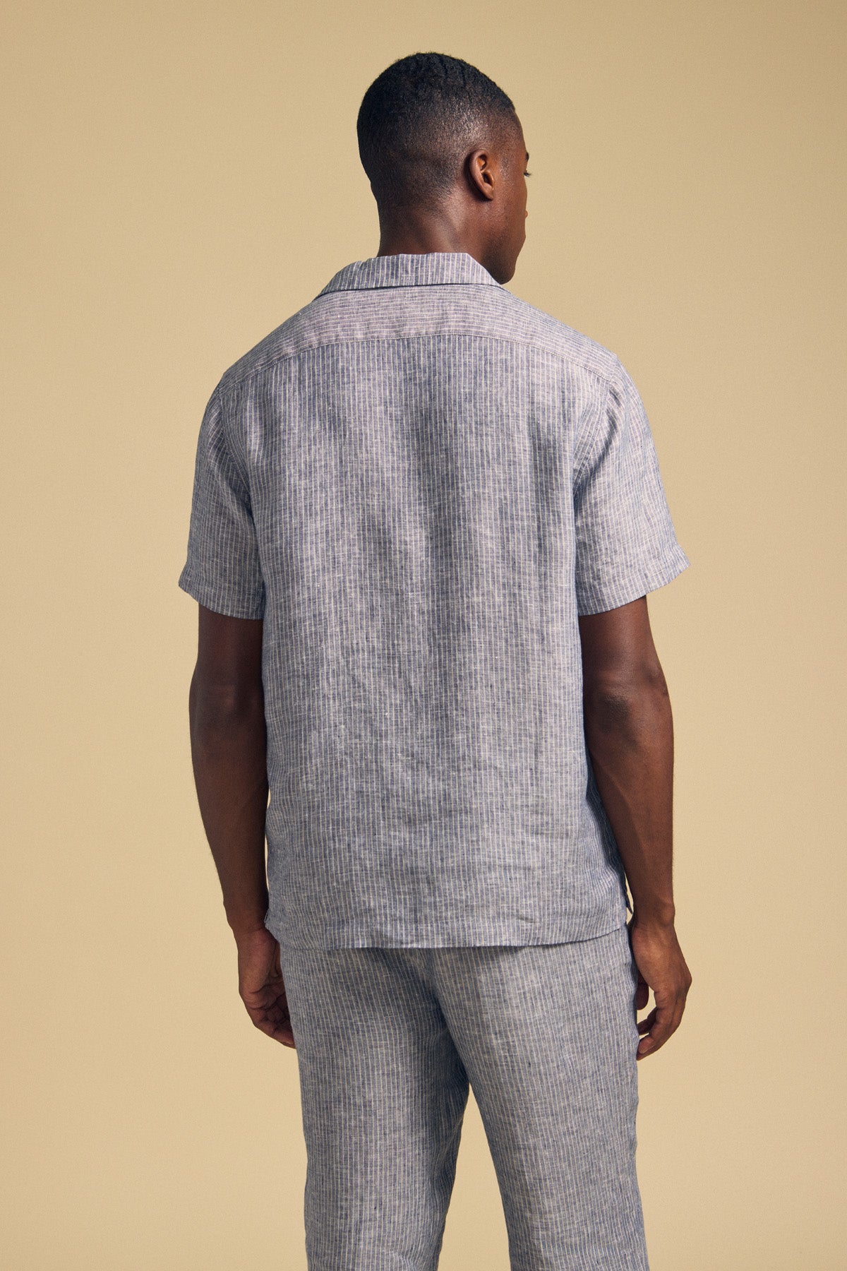 A man faces away, wearing the JULIUS LINEN SHIRT by Velvet by Graham & Spencer—a short-sleeve, camp collar, light gray striped button-up—paired with matching pants against a beige background.-41548474482881