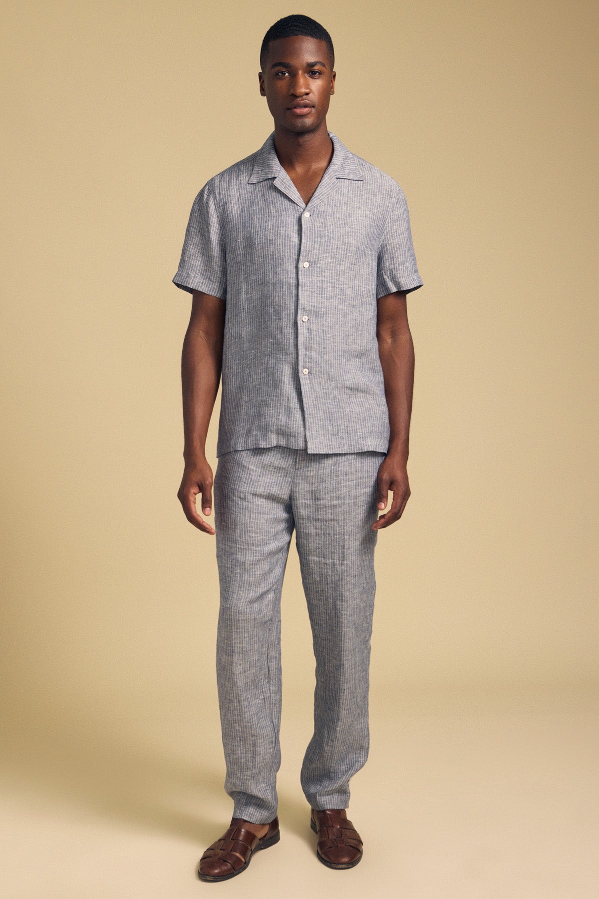 A man stands against a beige background wearing Velvet by Graham & Spencer’s JAE LINEN PANT in light blue and gray with an elastic waistband, paired with a matching short-sleeve shirt and brown sandals.-41548474581185