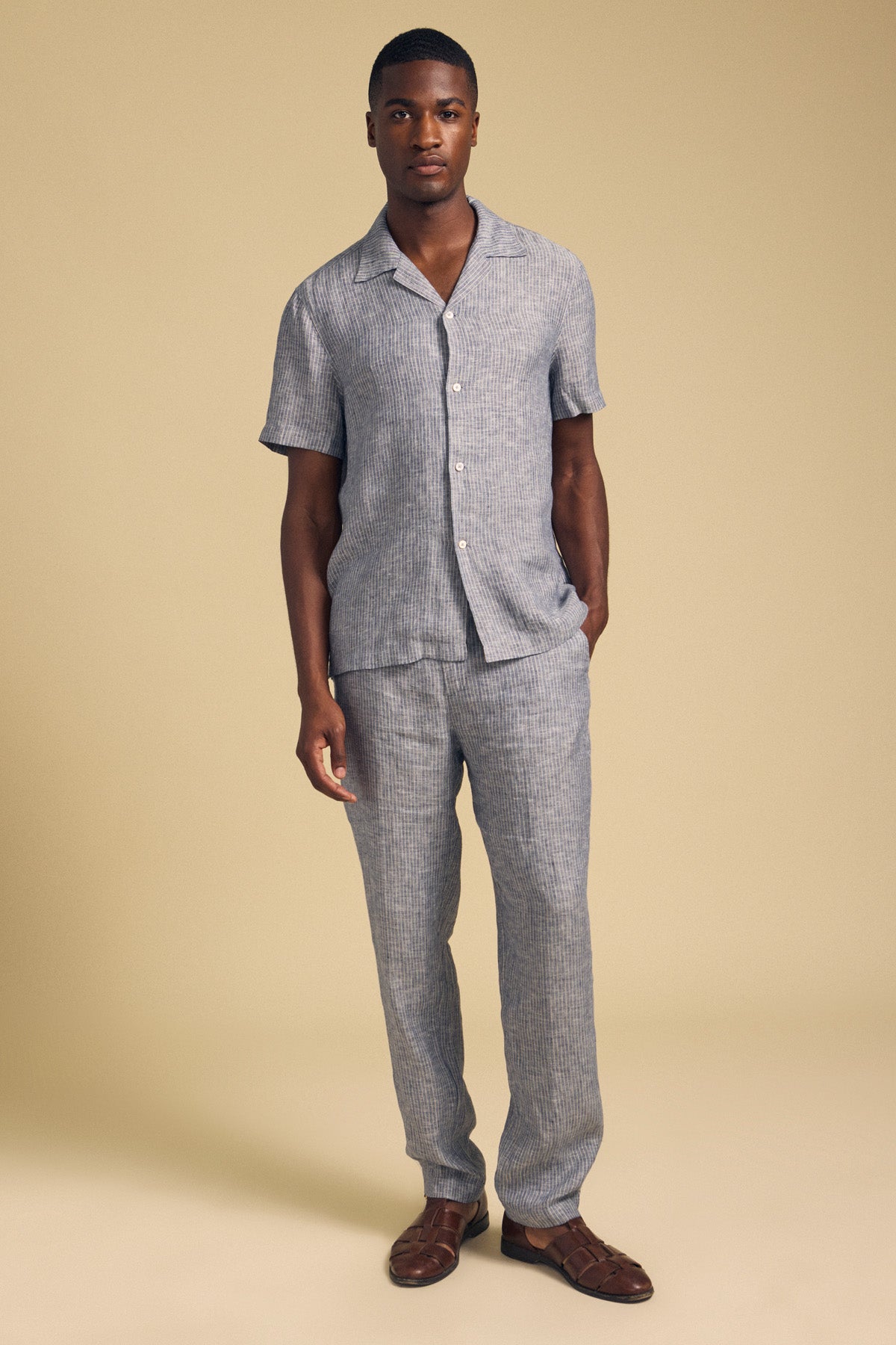 A man stands against a beige background, wearing a short-sleeve, button-up gray striped shirt and the Velvet by Graham & Spencer JAE LINEN PANT—straight-leg striped linen pants with an elastic waistband—paired with brown woven sandals.