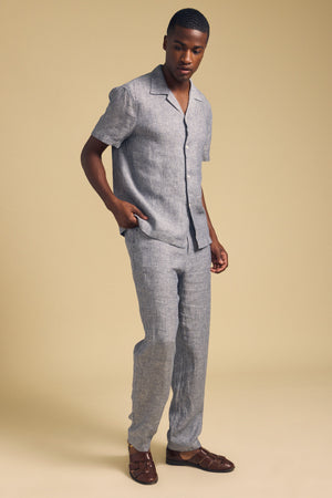 A man stands against a beige background wearing a short-sleeve, light grey striped shirt, JAE LINEN PANT by Velvet by Graham & Spencer—straight-leg, striped linen pants with an elastic waistband—and brown woven sandals.