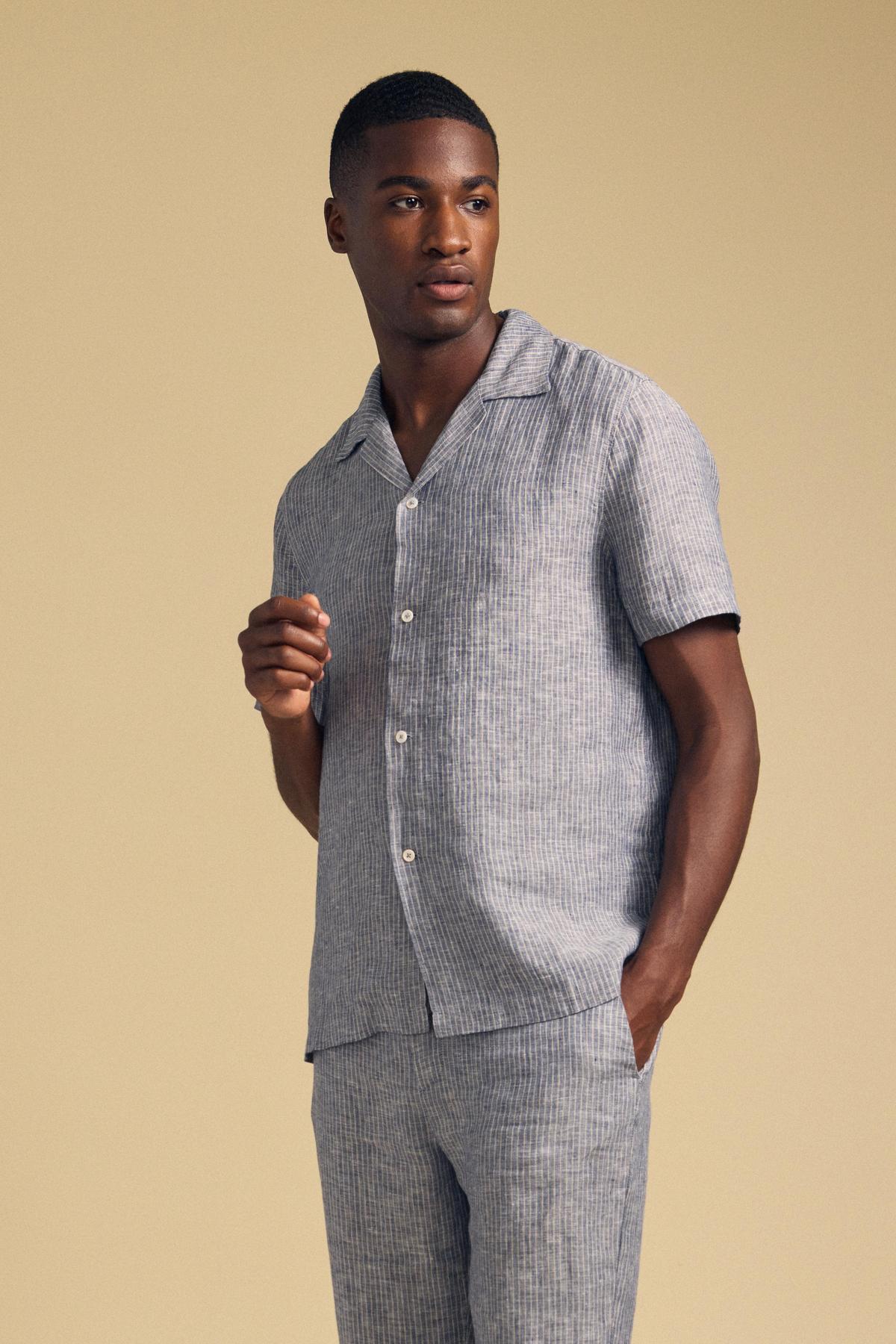 A man wears the JULIUS LINEN SHIRT by Velvet by Graham & Spencer— a short-sleeve, striped camp-collar button-up— with matching pants, standing against a plain beige background.