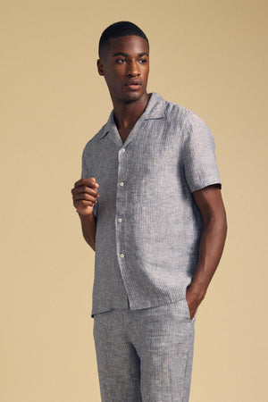 A man wears the JULIUS LINEN SHIRT by Velvet by Graham & Spencer— a short-sleeve, striped camp-collar button-up— with matching pants, standing against a plain beige background.
