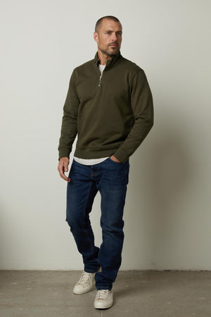 A versatile man wearing a green Velvet by Graham & Spencer BANNING QUARTER-ZIP SWEATSHIRT and jeans standing in front of a wall.