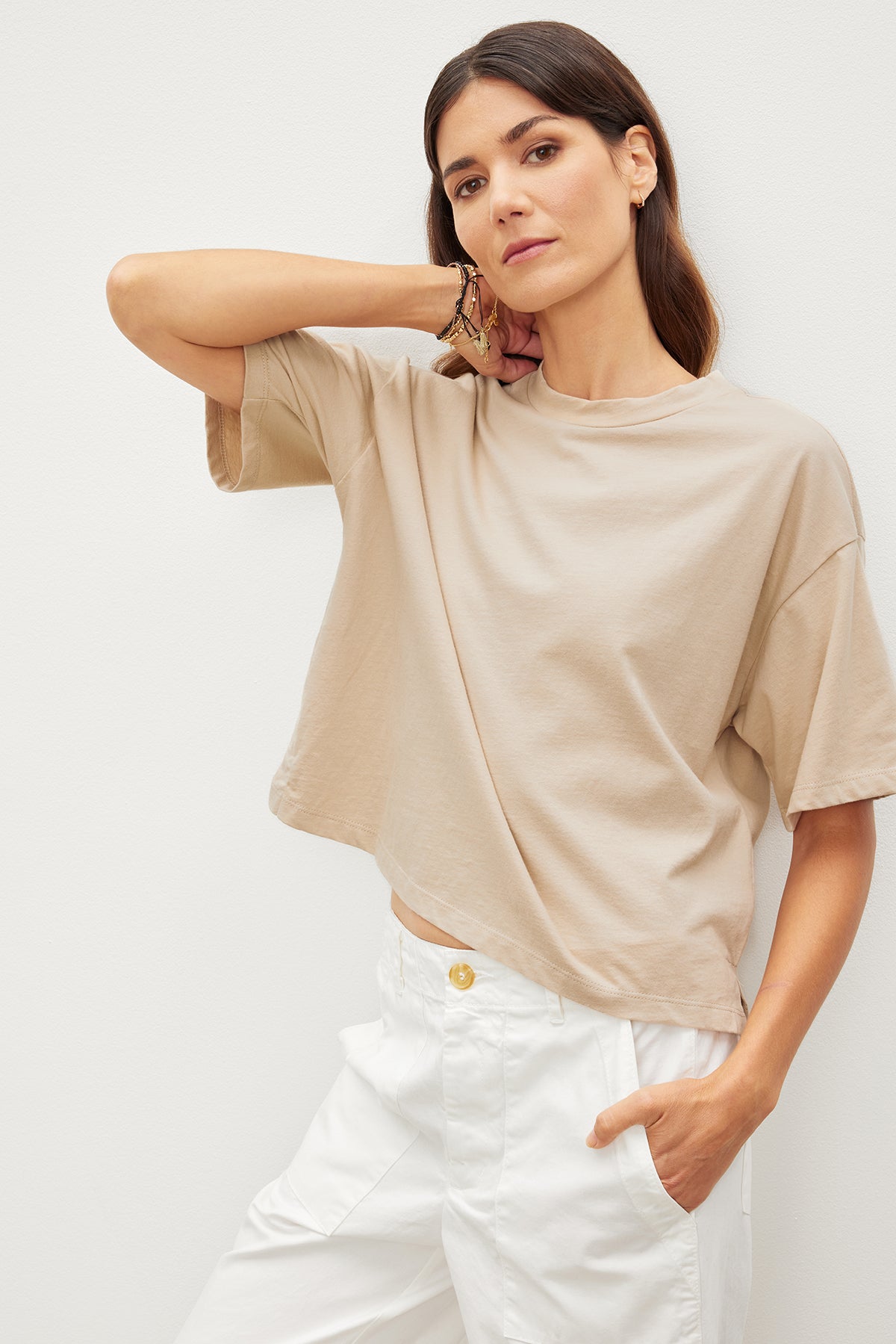   The model is wearing a relaxed beige CLARAH CREW NECK TEE crafted from sueded jersey fabric, paired with stylish white pants by Velvet by Graham & Spencer. 