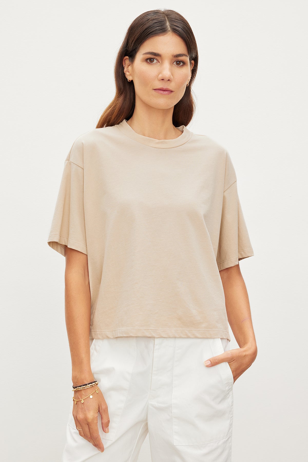   The model is wearing a Velvet by Graham & Spencer CLARAH CREW NECK TEE, a relaxed beige t-shirt made of sueded jersey fabric, and styled with white pants. 