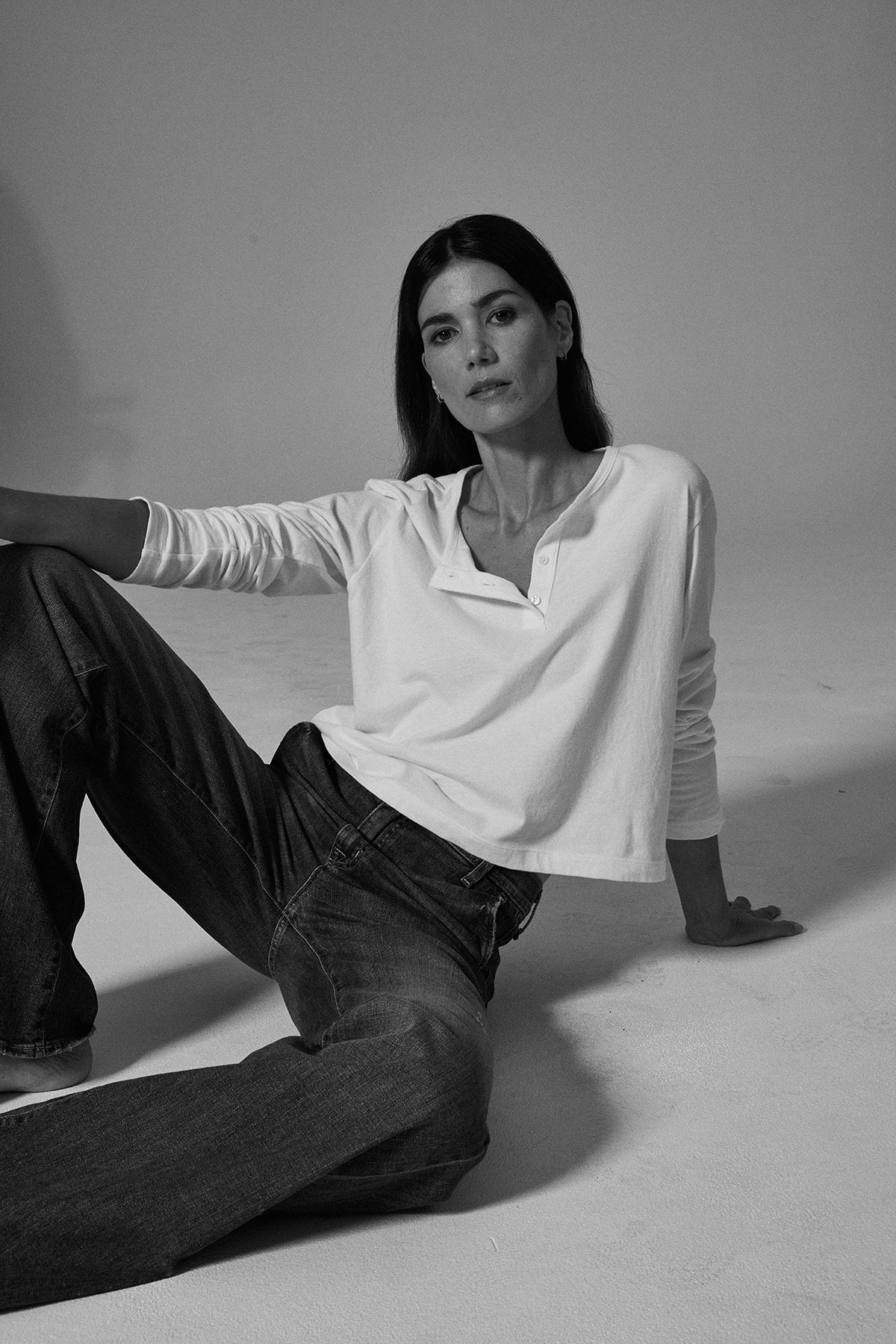   A woman sitting on the floor in DELIAH CROPPED HENLEY jeans made of sueded jersey cotton, along with a white shirt from Velvet by Graham & Spencer, featuring a cropped silhouette. 