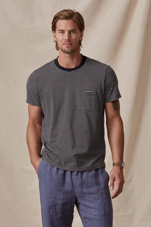 A man in a CHAZZ TEE from Velvet by Graham & Spencer stands against a beige backdrop, looking directly at the camera with a slight smile.