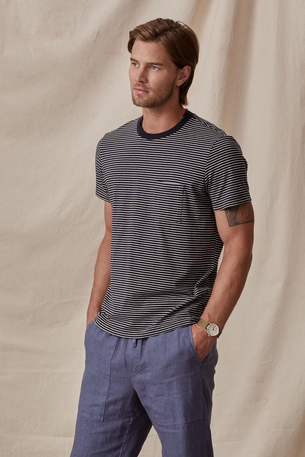 A man wearing a Velvet by Graham & Spencer CHAZZ TEE and blue trousers stands confidently, hands in pockets, against a neutral backdrop.-36732513026241