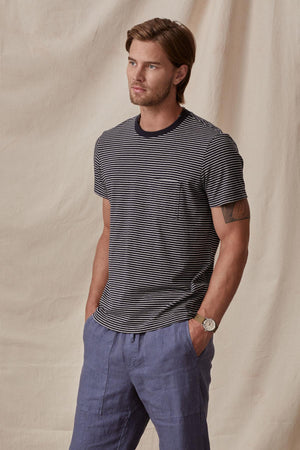 A man wearing a Velvet by Graham & Spencer CHAZZ TEE and blue trousers stands confidently, hands in pockets, against a neutral backdrop.