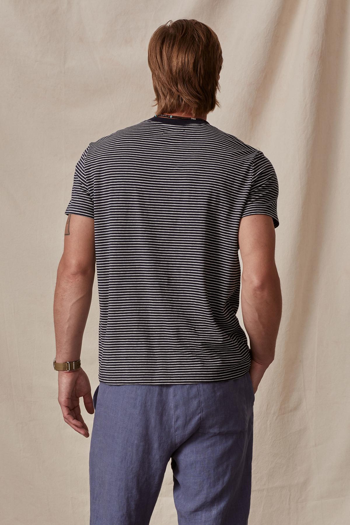 Man standing with his back to the camera, wearing a Velvet by Graham & Spencer CHAZZ TEE and blue trousers, in front of a beige backdrop.-36732513059009