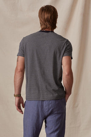 Man standing with his back to the camera, wearing a Velvet by Graham & Spencer CHAZZ TEE and blue trousers, in front of a beige backdrop.