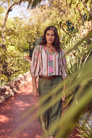 Woman walking down garden path wearing Beth Boho Top in multi colored jacquard print tucked into Dru pant in basil green with brown belt and necklace 