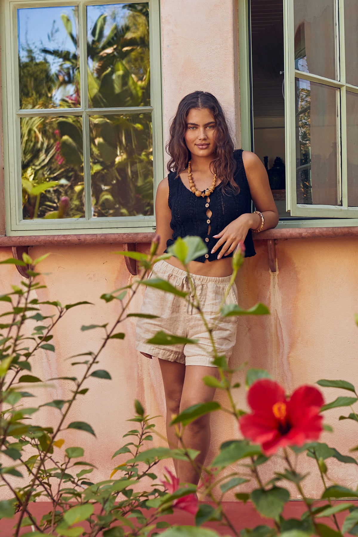   Woman standing outdoors leaning against wall of house with windows wearing Layla Crochet Stitch Tank Top in black with Tammy short in sand front with hibiscus in foreground 
