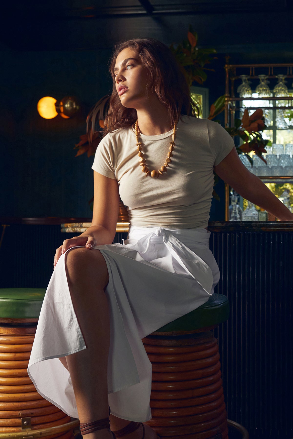 Women sitting on bar stool wearing Leena Skirt with tie in front white with Nina Tee in bisque tucked, chunky necklace and legs crossed to show skirt opening-26479438004417