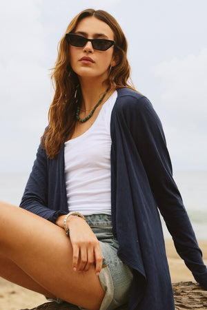 A woman wearing a Velvet by Graham & Spencer CHAMPAGNE OPEN CARDIGAN and denim shorts sitting on a rock.