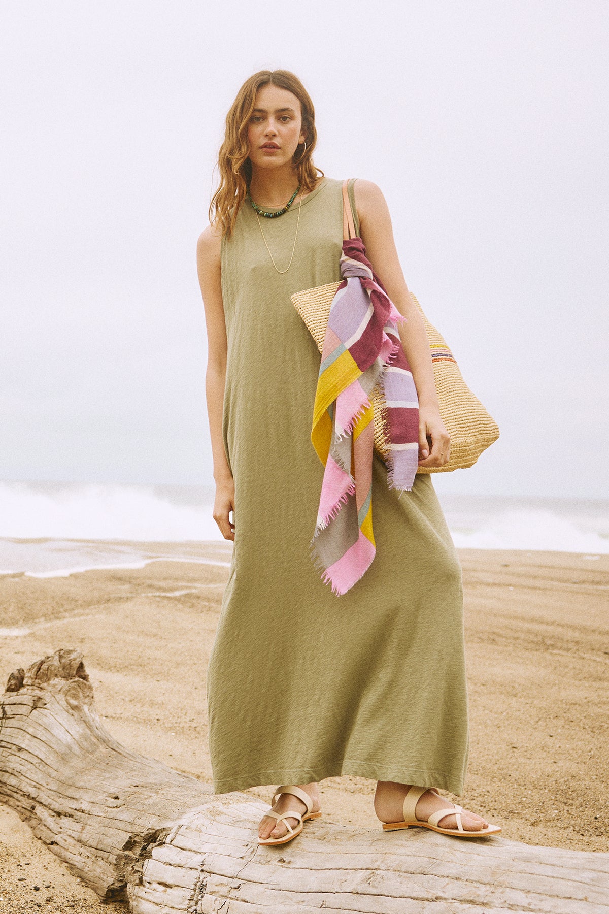   A woman in a axe green EDITH SLEEVELESS MAXI DRESS by Velvet by Graham & Spencer standing on long on the beach with a wicker basket. 