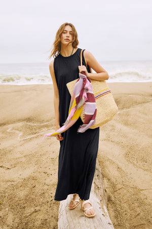 A woman in a black EDITH SLEEVELESS MAXI DRESS by Velvet by Graham & Spencer standing on the beach with a wicker basket.