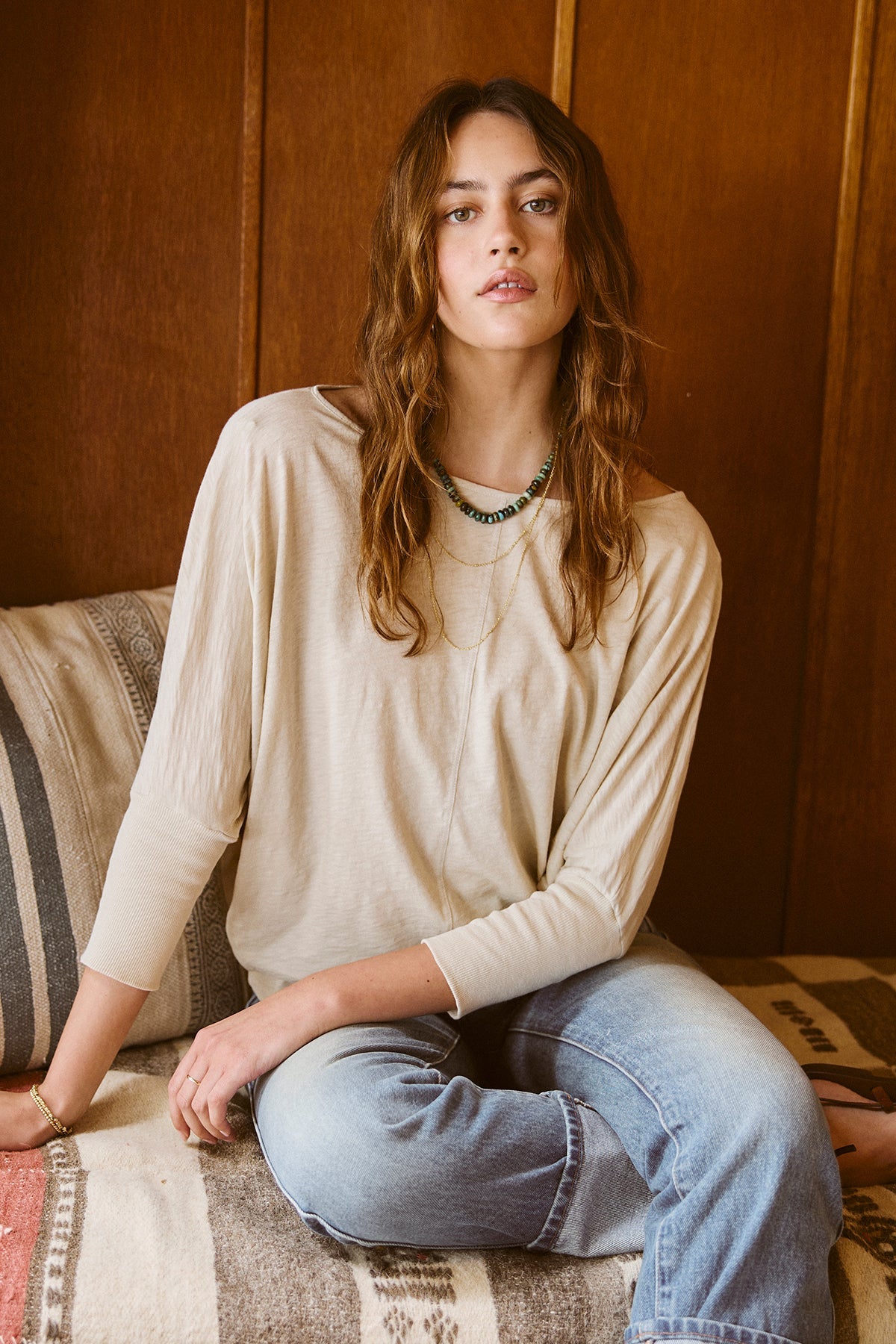   A woman sitting on a couch wearing JOSS DOLMAN SLEEVE TEE by Velvet by Graham & Spencer, jeans, and a beaded necklace 