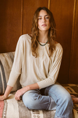 A woman sitting on a couch wearing JOSS DOLMAN SLEEVE TEE by Velvet by Graham & Spencer, jeans, and a beaded necklace