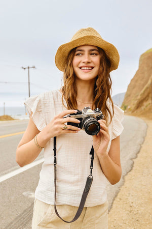 A young woman holding a REMI RUFFLE SLEEVE TOP camera on the side of the road. (Brand Name: Velvet by Graham & Spencer)
