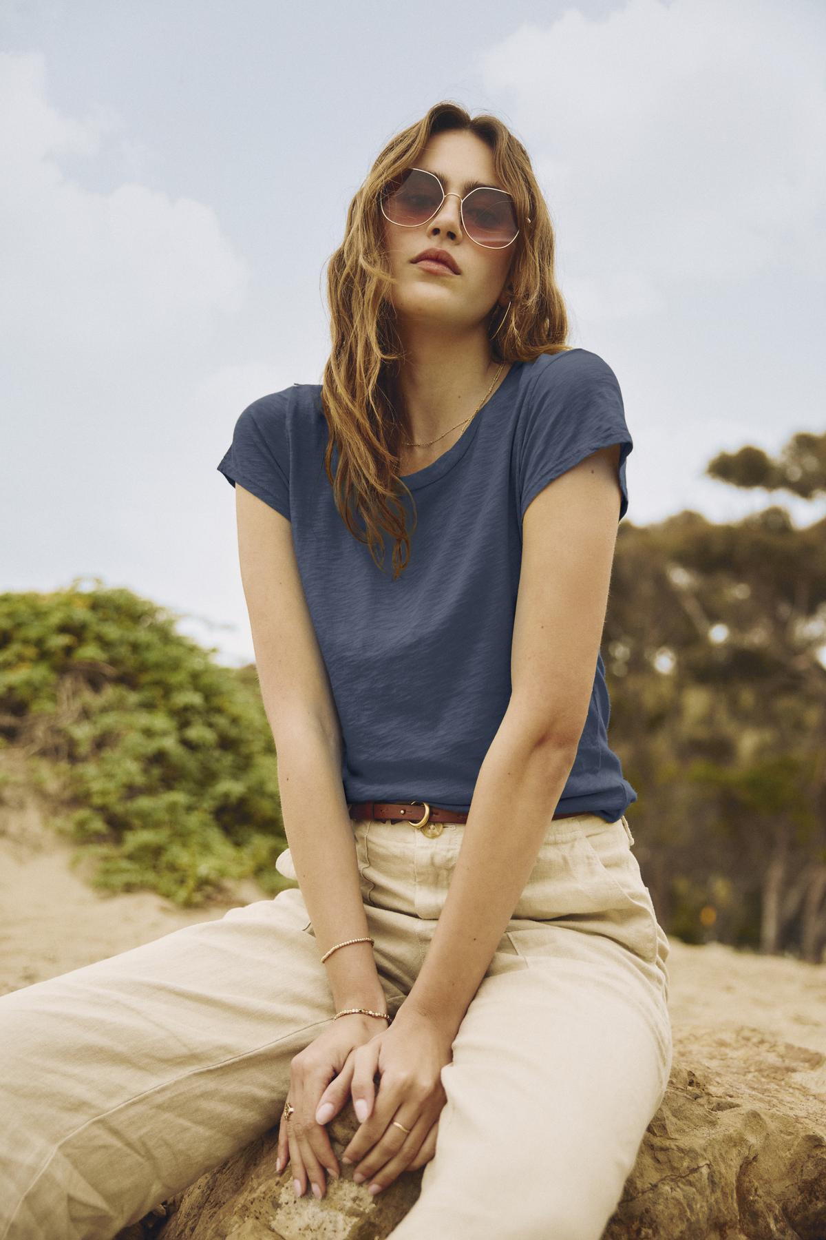 A person with wavy brown hair wearing sunglasses, a blue shirt made from premium cotton slub— the ODELIA TEE by Velvet by Graham & Spencer, a true wardrobe essential— and light-colored pants sits outdoors with a natural background.-37250157478081