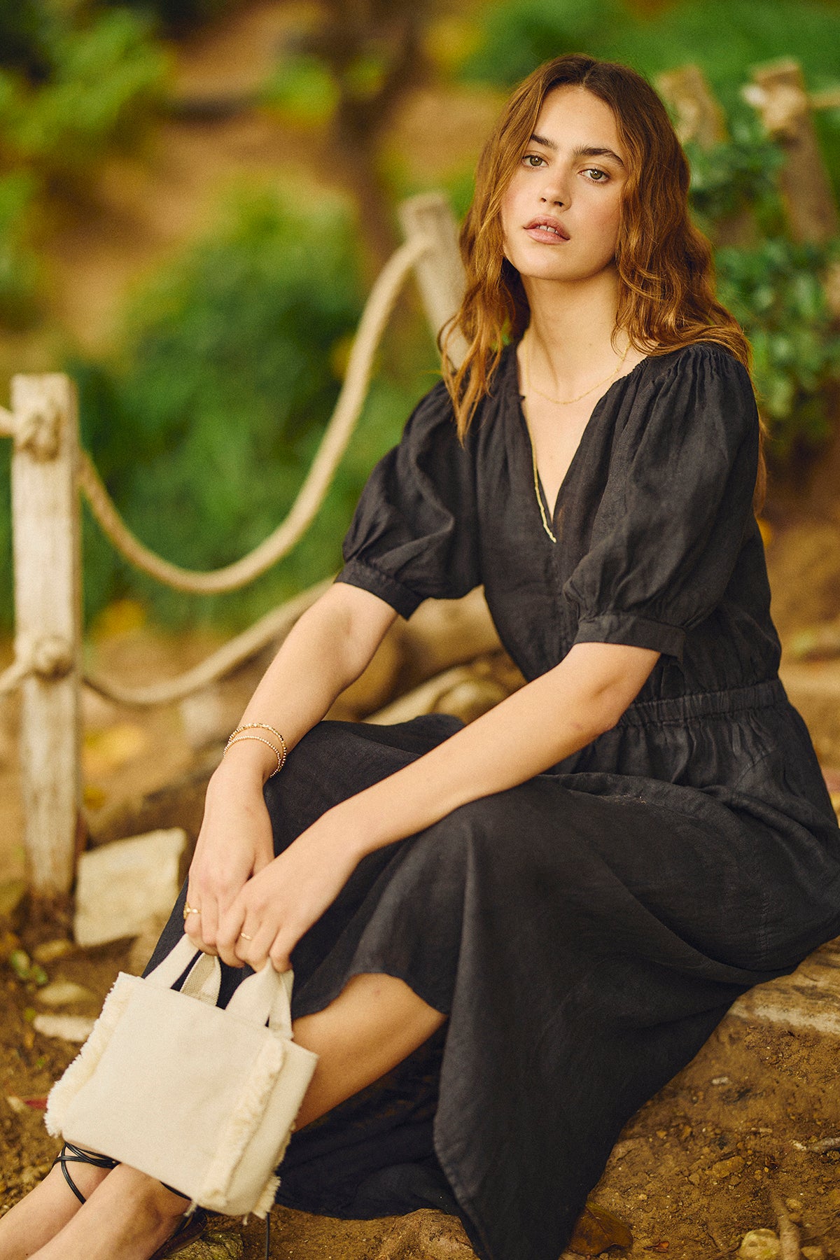 A woman in a black WHITNEY LINEN CUT OUT DRESS by Velvet by Graham & Spencer sits on a rock with the Min Launch Canvas Bag.-26671391375553