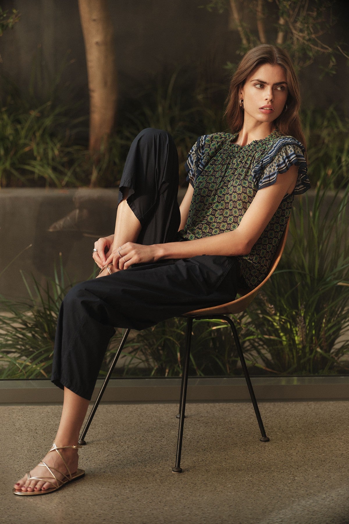A woman sits sideways on a chair, wearing the DAHLIA TOP by Velvet by Graham & Spencer with ruffle sleeves, paired with dark pants and sandals, surrounded by greenery and trees.-41526972645569