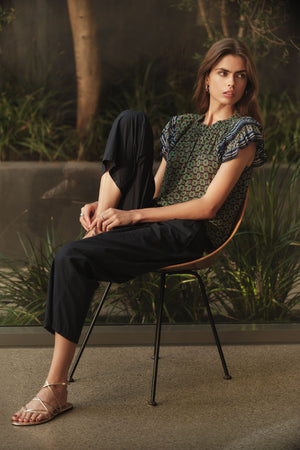 A woman sits sideways on a chair, wearing the DAHLIA TOP by Velvet by Graham & Spencer with ruffle sleeves, paired with dark pants and sandals, surrounded by greenery and trees.