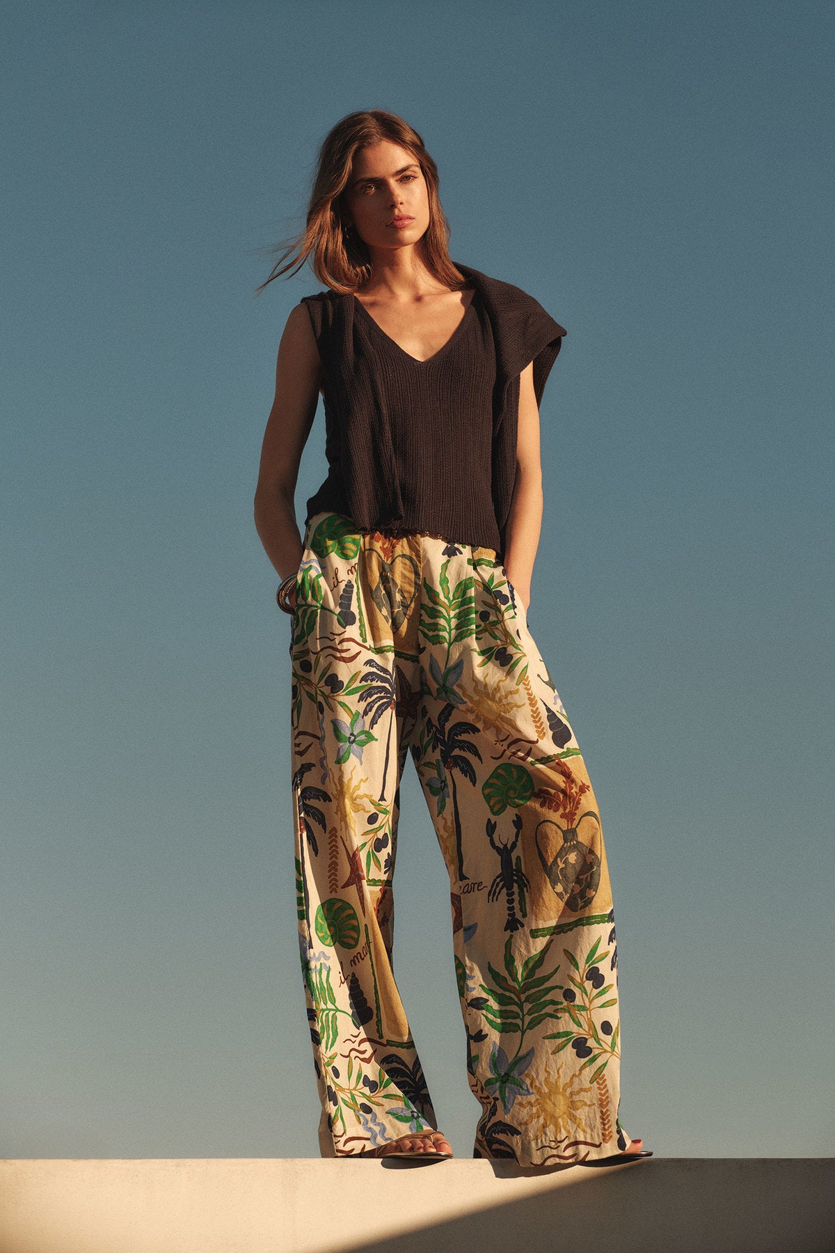 Woman standing outdoors in a black sleeveless top and the TRIXIE PANT by Velvet by Graham & Spencer, featuring a colorful botanical print and wide-leg silhouette, against a clear blue sky.-41527551230145