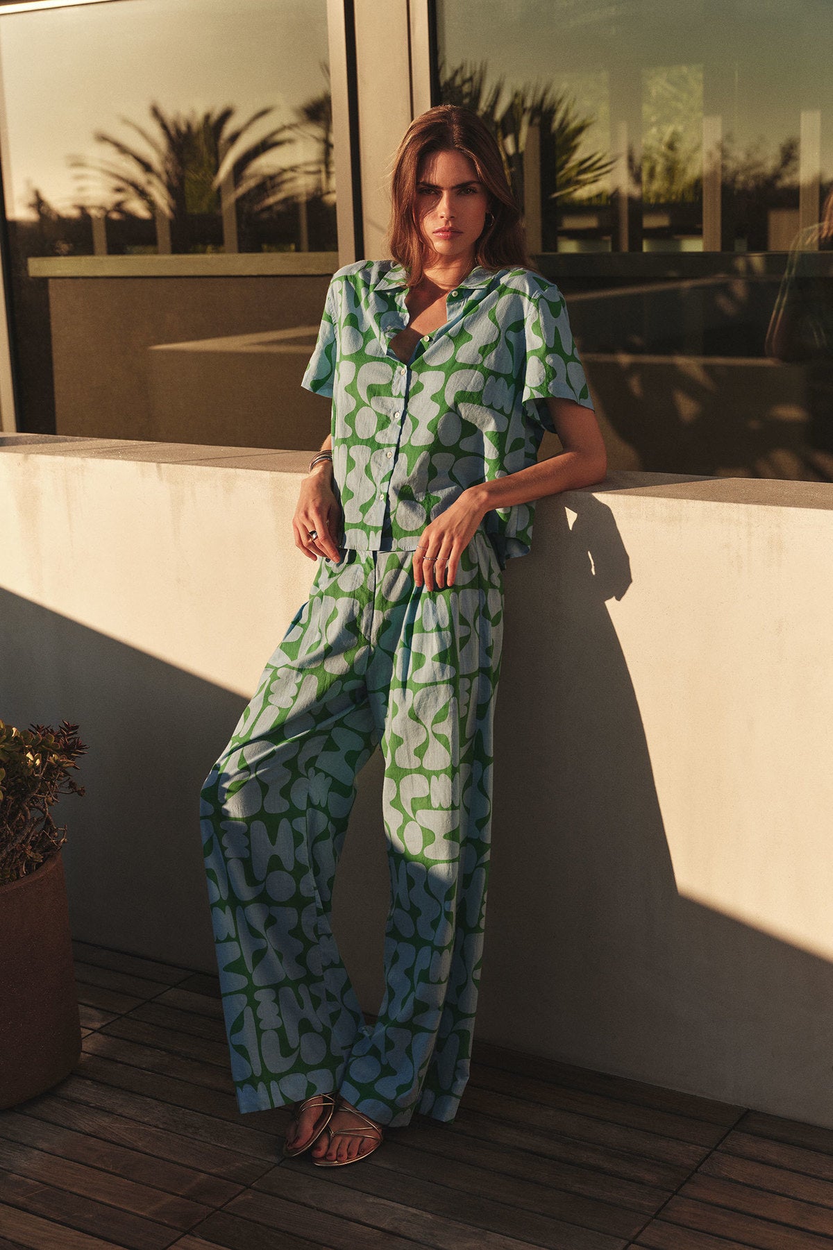 A woman stands on a patio in sunlight, wearing Velvet by Graham & Spencer’s TRIXIE PANT—a pair of resort-inspired, green and blue patterned cotton pants—styled with a matching short-sleeve blouse.