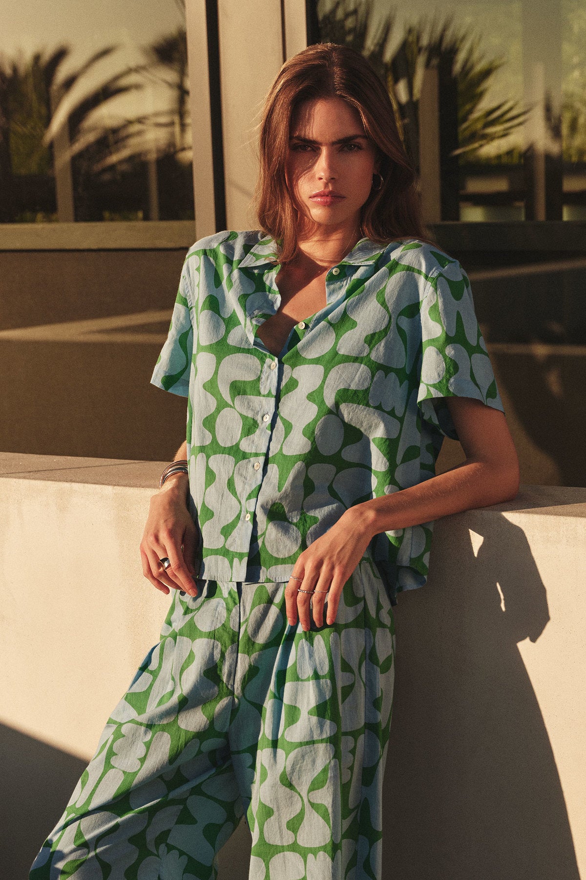 A woman stands outdoors against a sunlit wall, wearing the MARINE SHIRT by Velvet by Graham & Spencer in a blue and green Lido-printed cotton abstract pattern with matching pants.-41679984066753