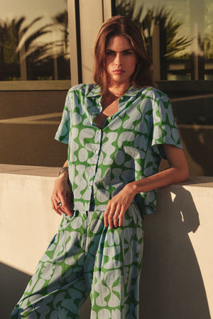 A woman stands outdoors against a sunlit wall, wearing the MARINE SHIRT by Velvet by Graham & Spencer in a blue and green Lido-printed cotton abstract pattern with matching pants.