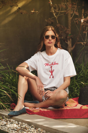 A woman wearing sunglasses, shorts, and the Velvet by Graham & Spencer SAMMY TEE sits on a patterned towel outdoors near plants and rocks.