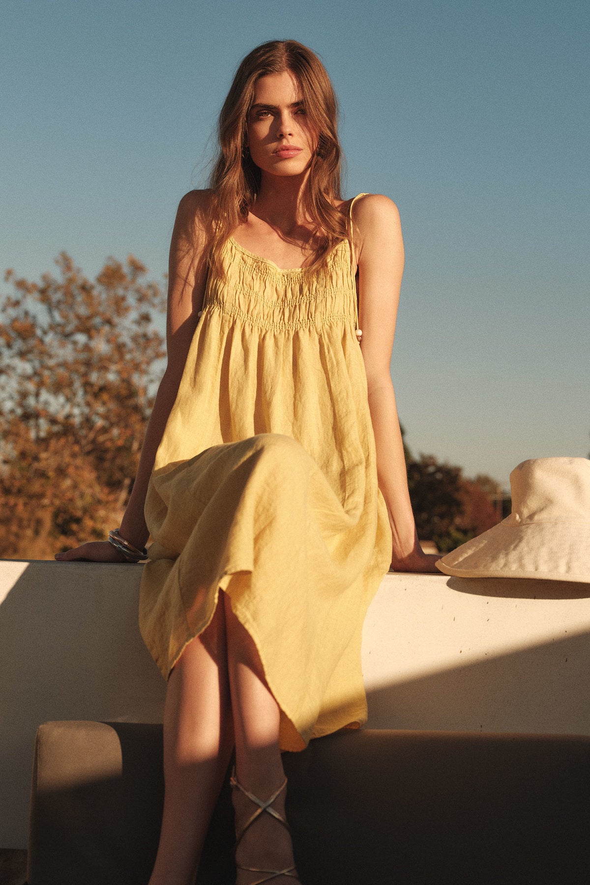 A woman in the TERESA LINEN DRESS by Velvet by Graham & Spencer, a yellow linen midi with a smocked bust, sits on a sunlit outdoor ledge with a white hat beside her.-41617591468225