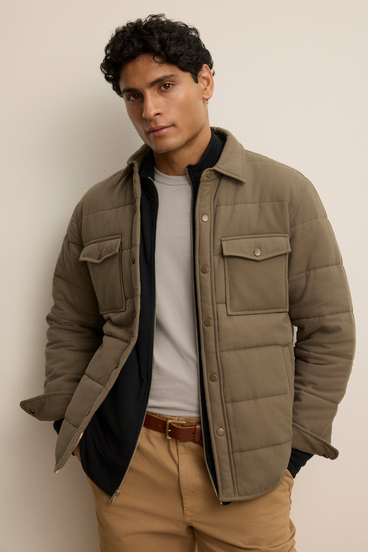   A man stands against a light wall wearing the SUMMIT JACKET by Velvet by Graham & Spencer—a tan quilted cotton jacket with snap buttons—over a light gray shirt and tan pants with a brown belt. 