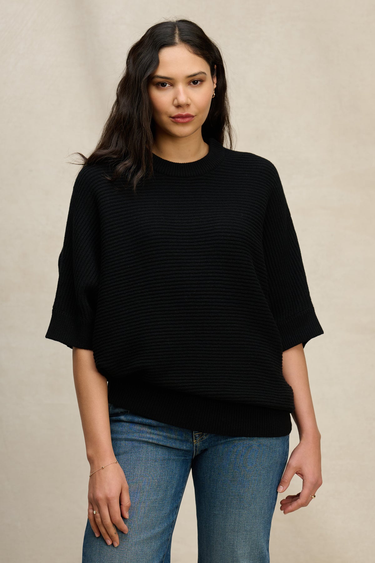  A woman with long dark hair wears the COCO SWEATER by Velvet by Graham & Spencer, a black wool blend crew neck, paired with blue jeans against a beige background. 