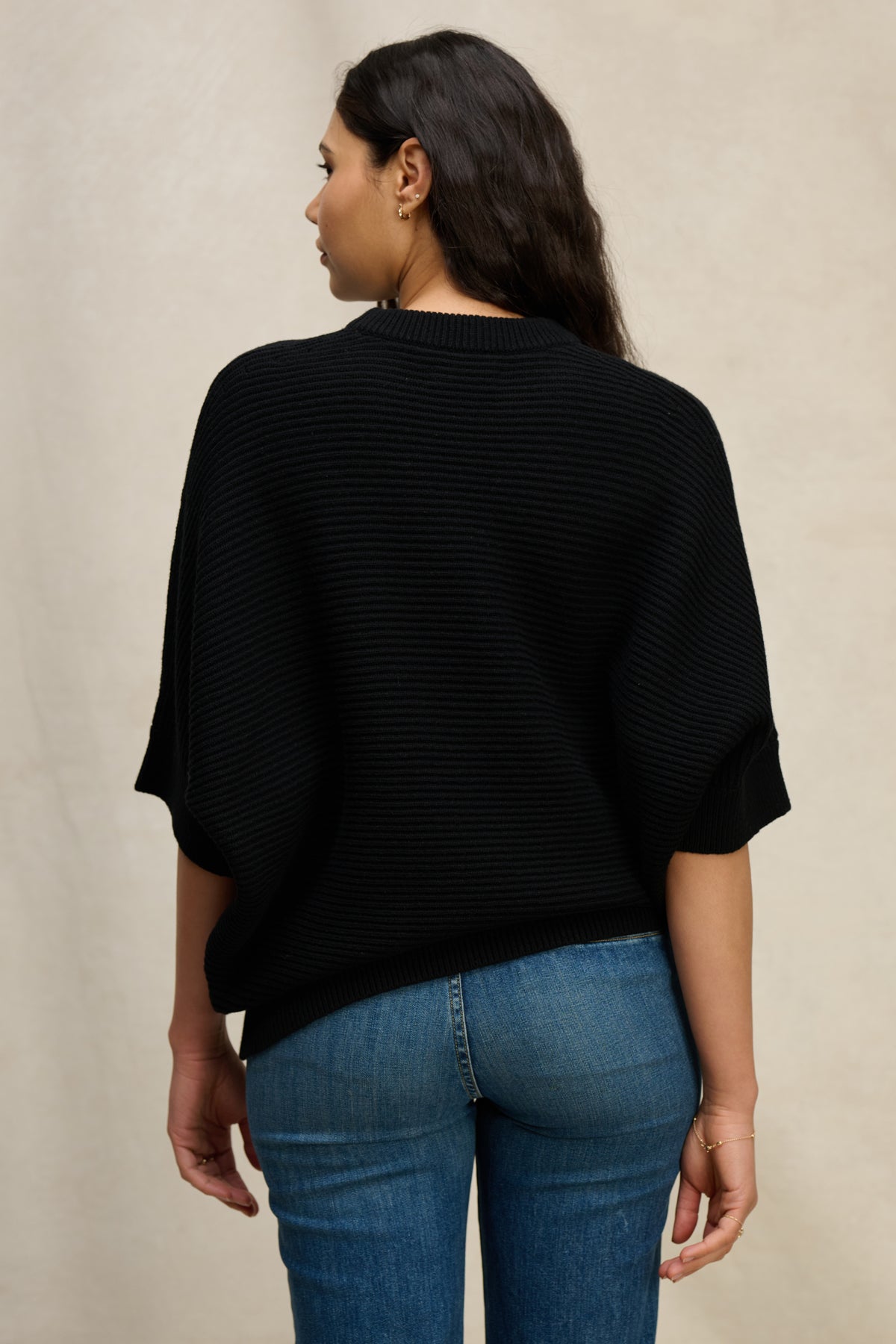  A woman with long dark hair is seen from behind wearing the Velvet by Graham & Spencer COCO SWEATER—a loose black wool blend in a cocoon silhouette—paired with blue jeans against a neutral background. 