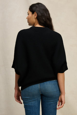 A woman with long dark hair is seen from behind wearing the Velvet by Graham & Spencer COCO SWEATER—a loose black wool blend in a cocoon silhouette—paired with blue jeans against a neutral background.