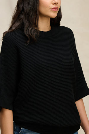 A woman wears the COCO SWEATER by Velvet by Graham & Spencer—a black, ribbed, wool blend crewneck—against a neutral background. Only her torso and lower face are visible.