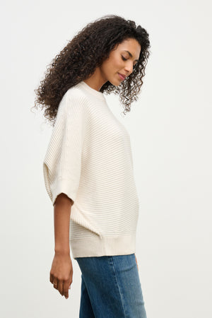 A woman with curly hair wears the cream-colored COCO SWEATER by Velvet by Graham & Spencer, featuring a crew neckline, paired with blue jeans and standing in profile against a plain white background.