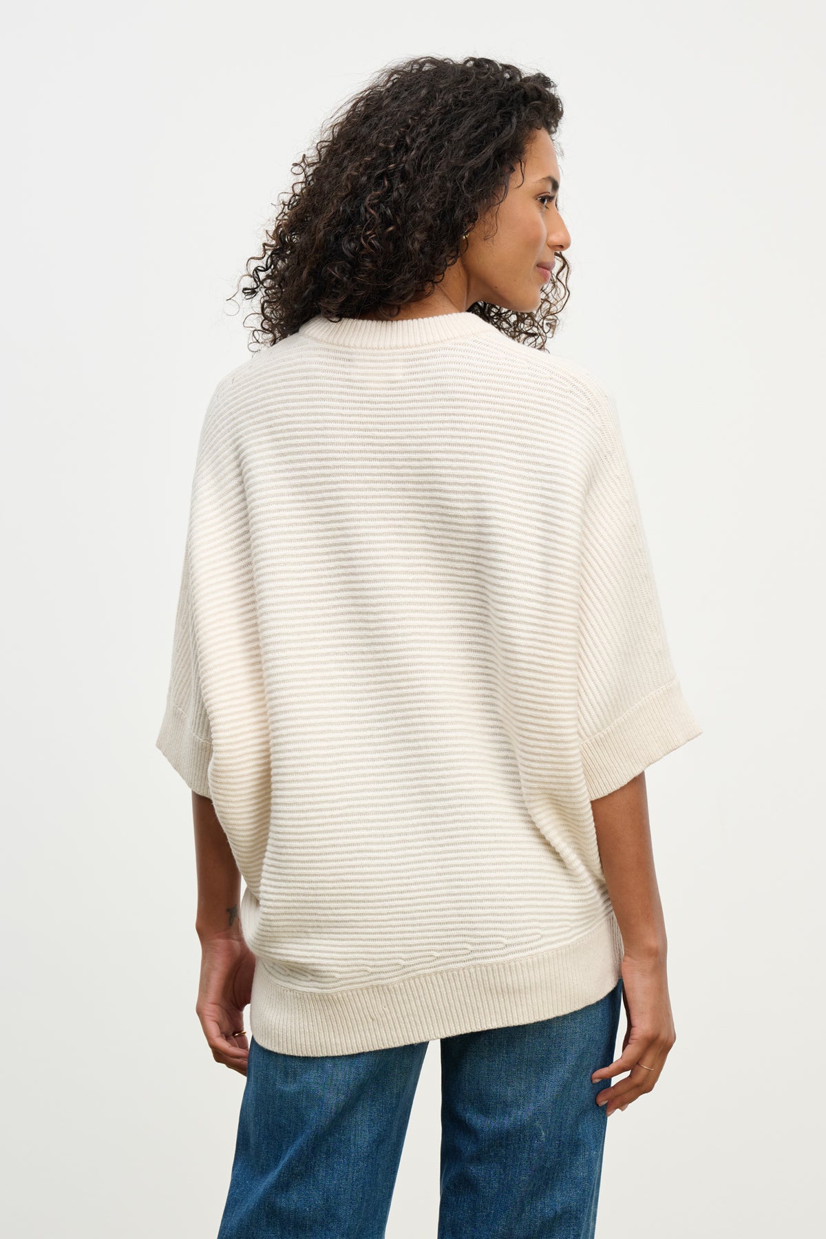 A woman with curly hair wears the COCO SWEATER by Velvet by Graham & Spencer—a loose, cream wool blend with a crew neckline—paired with blue jeans, standing and facing away from the camera.-40227040657601