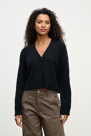 A person wears the ETNA CARDIGAN by Velvet by Graham & Spencer—a black wool blend V-neck sweater—paired with brown pants, standing against a plain light background, making it the ideal layering piece for any outfit.