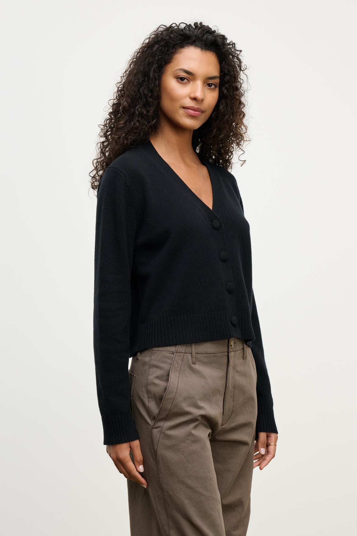 A woman with curly hair wears the ETNA CARDIGAN by Velvet by Graham & Spencer—a black wool blend V-neck cardigan—paired with khaki pants, standing against a plain light background.
