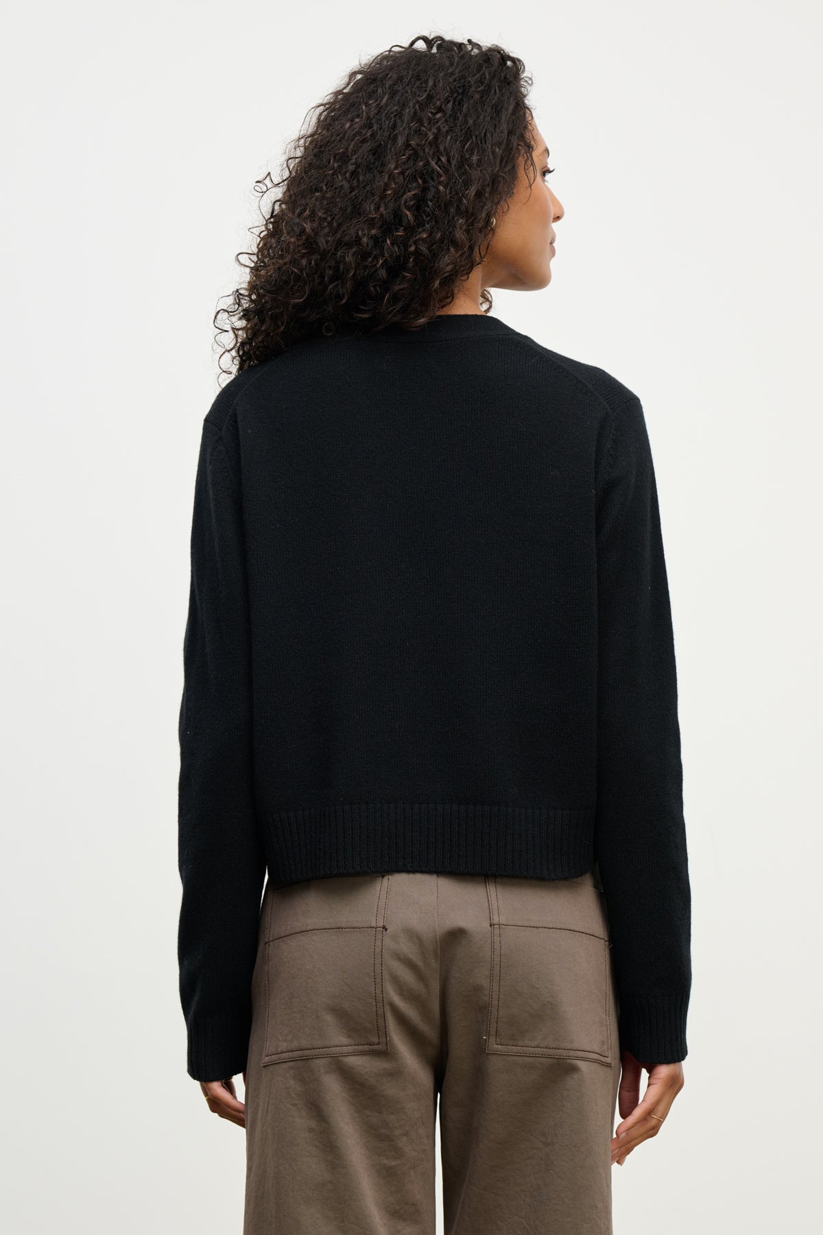 A person with curly hair is seen from behind, wearing the ETNA CARDIGAN by Velvet by Graham & Spencer in black over brown pants, set against a plain light background.