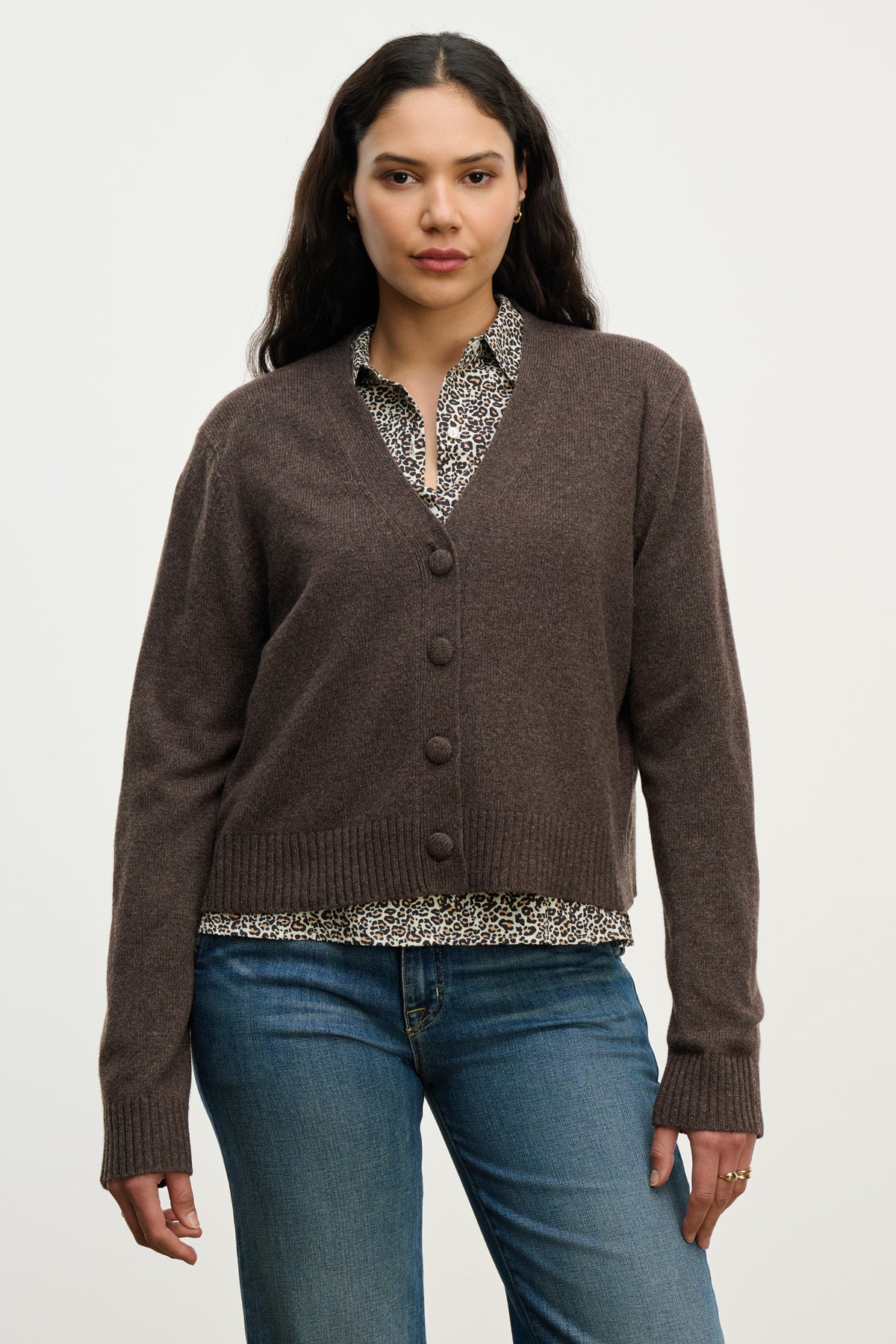   A woman wears the ETNA CARDIGAN by Velvet by Graham & Spencer—a brown wool blend V-neck cardigan—over a patterned blouse and blue jeans, standing against a plain light background. 