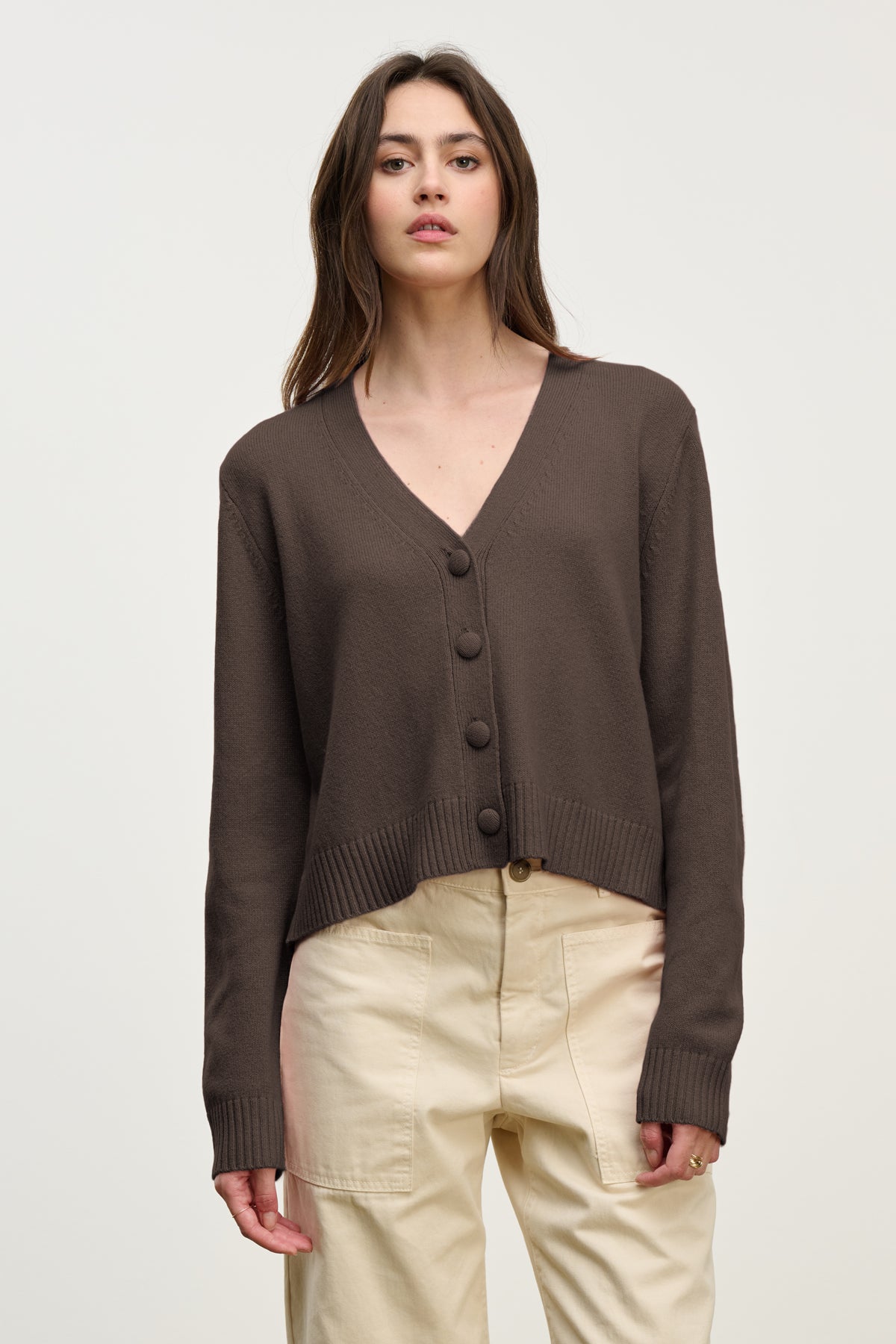   A woman wears the Velvet by Graham & Spencer ETNA CARDIGAN, a brown wool blend V-neck, paired with high-waisted beige pants against a plain light background. 