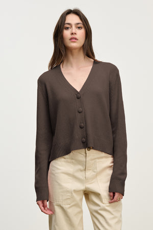 A woman wears the Velvet by Graham & Spencer ETNA CARDIGAN, a brown wool blend V-neck, paired with high-waisted beige pants against a plain light background.