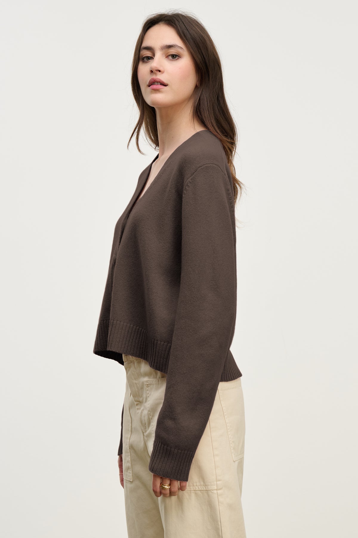 Woman in a side pose wears the ETNA CARDIGAN by Velvet by Graham & Spencer—a brown V-neck cardigan styled with beige pants against a light background, showcasing this versatile layering piece for any season.-39814881607873