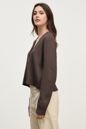 Woman in a side pose wears the ETNA CARDIGAN by Velvet by Graham & Spencer—a brown V-neck cardigan styled with beige pants against a light background, showcasing this versatile layering piece for any season.